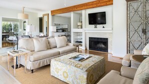 A living room with a sofa, ottoman, and coffee table, a fireplace, and a television mounted on the wall. - Kiawah Exclusives | 4973 Turtle Point Villa (Kiawah Island)