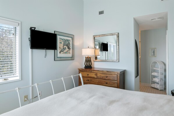 Un dormitorio con cama, cómoda de madera, mesita de noche con lámpara, televisor montado en la pared y ventana con persianas.