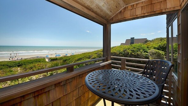 Un balcón de madera con una mesa y sillas de metal, con vistas a una playa donde hay gente y sombrillas.