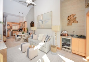 Una sala de estar moderna con un sofá, un reposapiés y una mesa de comedor con sillas. Se aprecia una cocina al fondo con un fregadero y armarios.