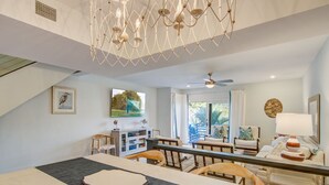 A dining area with a long table, chairs, a chandelier, a ceiling fan, and a view of the living room with a sofa and a lamp.