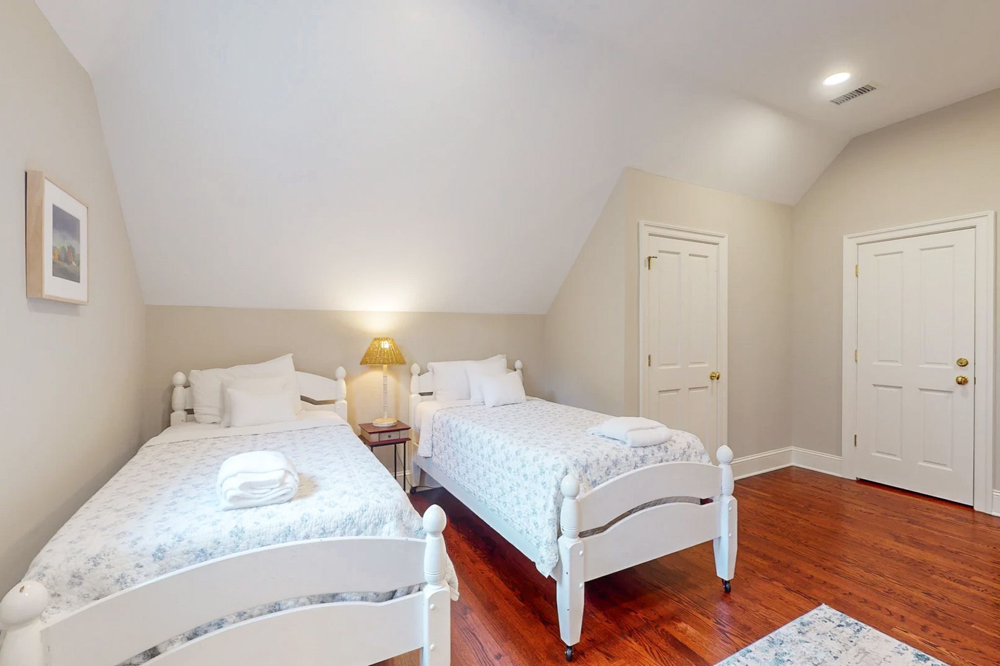 Un dormitorio con dos camas individuales, una mesita de noche con lámpara y una pequeña alfombra sobre el suelo de madera.
