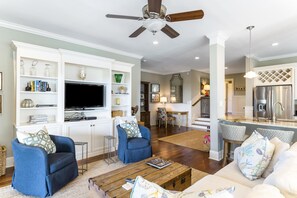 A spacious living room with a flat-screen TV, two blue armchairs, a wooden coffee table, and a white sofa.