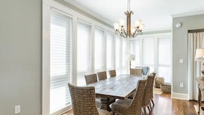 A dining room with a large wooden table surrounded by wicker chairs, a chandelier, and large windows with blinds.