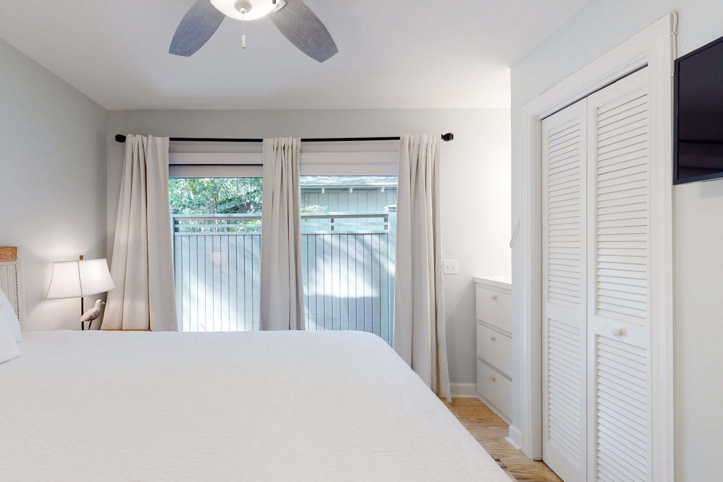 A bedroom with a bed, a ceiling fan, a window with curtains, a nightstand with a lamp, and a white wardrobe.