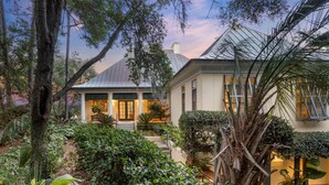 A house with a covered porch, surrounded by lush greenery and a large tree. - Kiawah Exclusives | 305 Ocean Oaks (Kiawah Island)