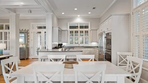 A spacious kitchen with white cabinetry, a central island, and a dining area with white chairs. - Kiawah Exclusives | 305 Ocean Oaks (Kiawah Island)