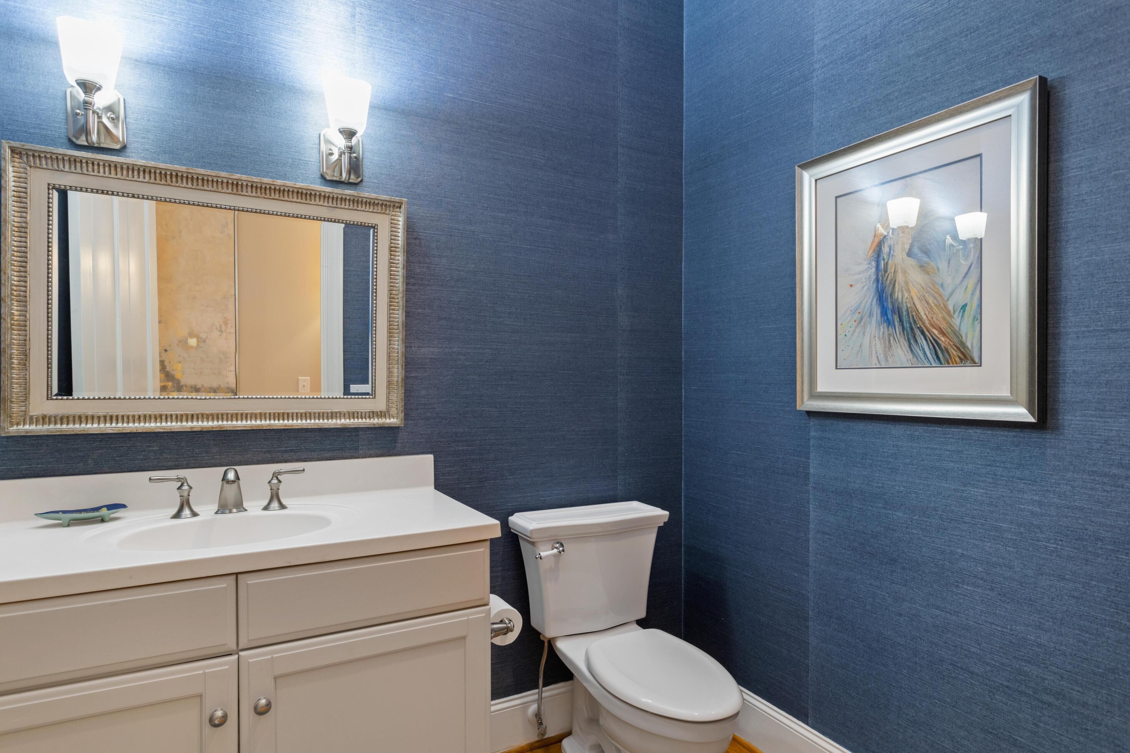 A bathroom with a white sink, two faucets, a toilet, a mirror with lights, and a framed painting on the wall.