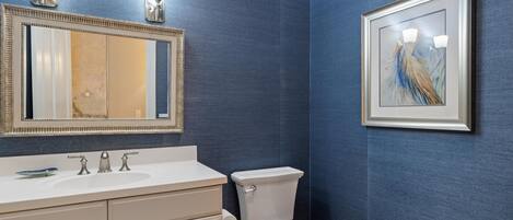 A bathroom with a white sink, two faucets, a toilet, a mirror with lights, and a framed painting on the wall.