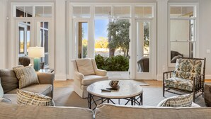 A spacious living room with a large window, a ceiling fan, and a round coffee table. - Kiawah Exclusives | 305 Ocean Oaks (Kiawah Island)