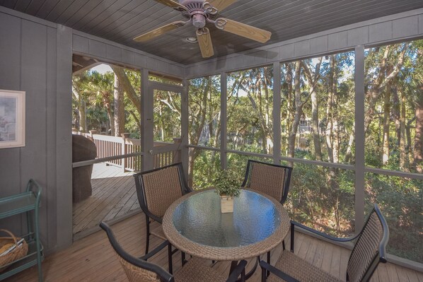 A covered outdoor seating area with a glass table and wicker chairs, overlooking a forested area.