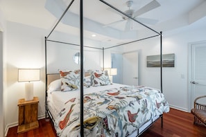 A four-poster canopy bed with a colorful bird-themed bedspread, a wooden nightstand with a lamp, and a ceiling fan.
