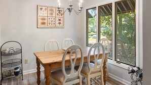 Un comedor con mesa de madera, sillas blancas, una araña, un cuadro en la pared y ventana con vista a vegetación.