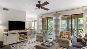 Amplia sala de estar con televisión de pantalla plana, asientos cómodos, ventilador de techo y ventanas grandes que ofrecen vistas al exterior.