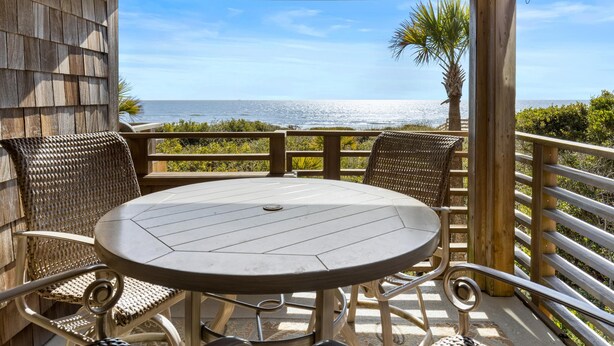 Une terrasse en bois avec une table ronde et des chaises en osier, donnant sur une plage bordée de palmiers et sur l’océan.