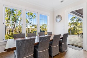 Un comedor con una mesa de madera y sillas de mimbre, ventanas amplias con vista a árboles, y un reloj en la pared.