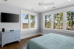 A bedroom with a bed, a TV, a dresser, and large windows with a view of trees and a beach.