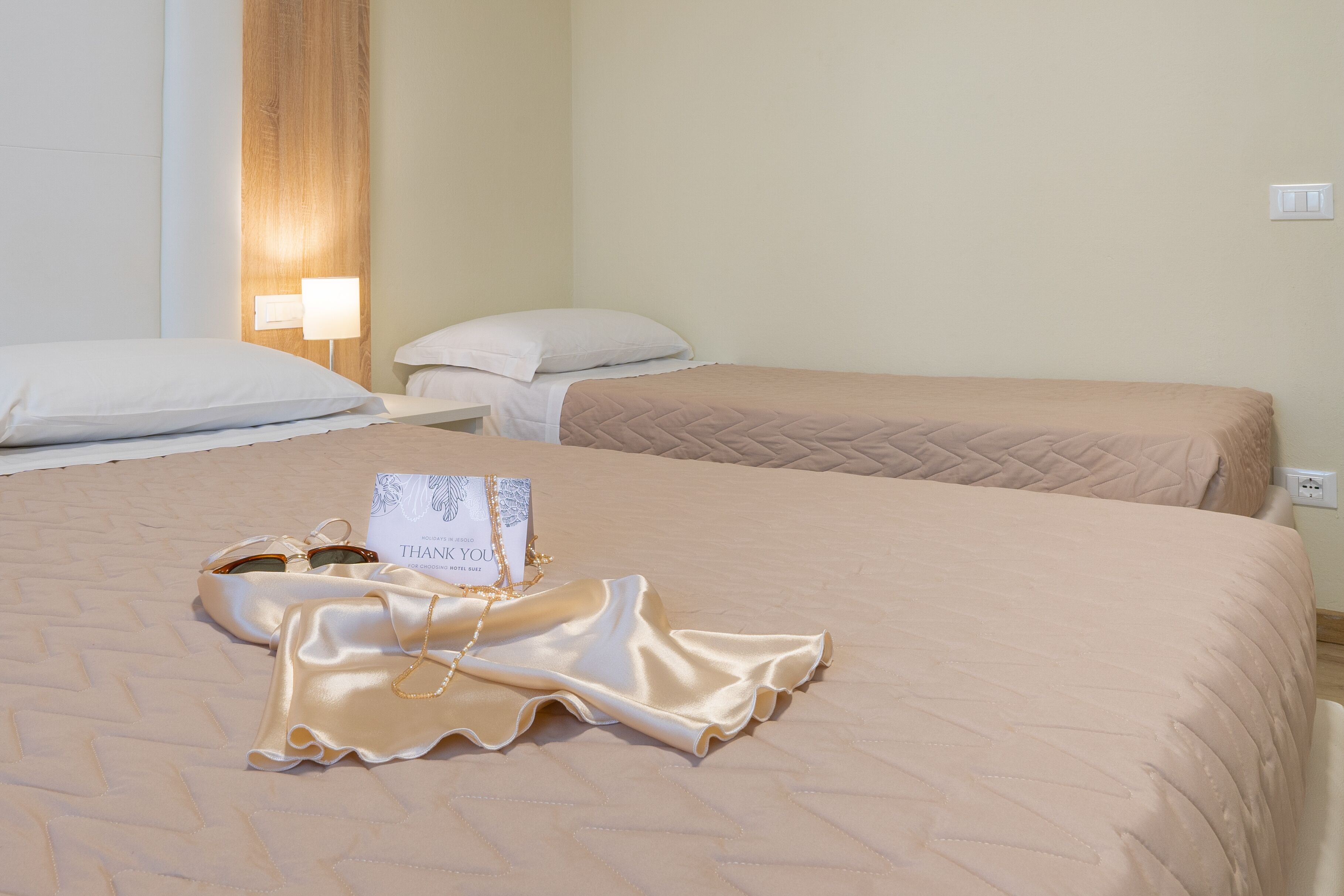 A hotel room with two beds, a bedside table, a lamp, and a message card on the bed.