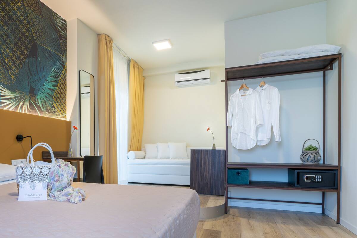 A hotel room with a bed, a bedside table with a lamp, a wardrobe with hanging clothes, and a wall-mounted air conditioner.