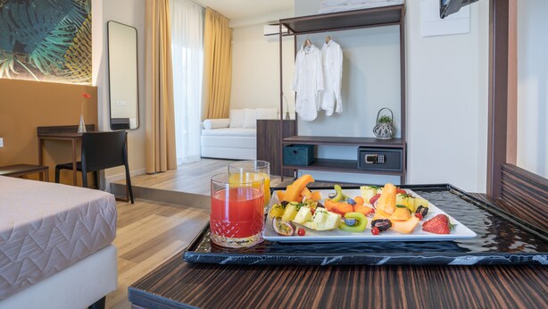 Ein Hotelzimmer mit Bett, Schreibtisch, Stuhl, Esstisch mit Obst und Saft sowie einem Kleiderständer.