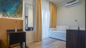 Ein modernes Hotelzimmer mit Holzboden, einer weißen Couch, einem dunklen Holztisch, einem Spiegel und einem großen Fenster mit Vorhängen.
