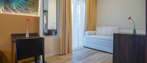 Habitació d'hotel moderna amb terra de fusta, un sofà blanc, escriptori de fusta fosca, mirall i una finestra gran amb cortines.