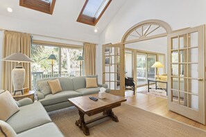 Un salon spacieux avec un canapé, une table basse et des lampes. Il y a une grande fenêtre et une porte qui donne sur un espace extérieur.