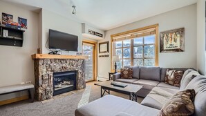 A living room with a stone fireplace, a television mounted on the wall, a sofa, and a window with a view of trees. - Beautiful Passage Point Condo.  Two blocks to American Eagle and American Flyer lifts. PP319 (Copper Mountain)