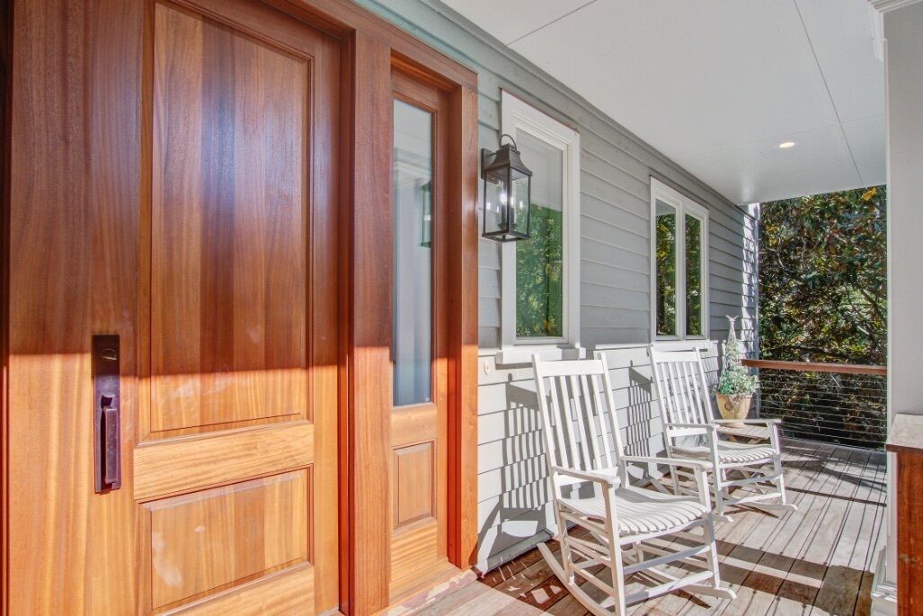 A wooden front door with a glass panel, white rocking chairs on a wooden deck, and a window with a view of greenery.