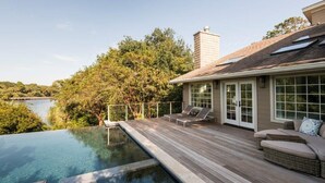 Un espace aménagé au bord de la piscine, avec une terrasse en bois, des chaises longues et une vue sur un lac entouré d’arbres.