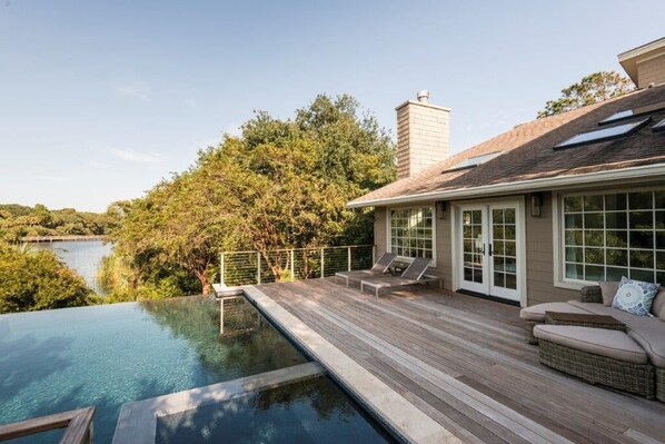A poolside area with a wooden deck, lounge chairs, and a view of a lake surrounded by trees.