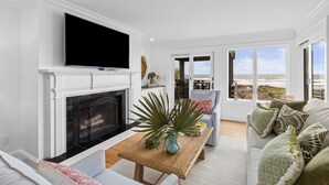 A living room with a fireplace, a television mounted above it, a dining area with a table and chairs, and a view of the beach through large windows. - Kiawah Exclusives | 4303 Windswept Villa (Kiawah Island)