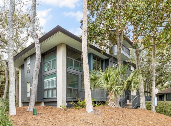 A modern two-story house with large windows, a flat roof, and a landscaped garden with trees and a palm plant.