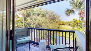 A balcony with a view of a golf course, a chair, a small table, and a decorative vase. - Kiawah Exclusives | 4942 Turtle Point Villa (Kiawah Island)