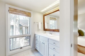 Une salle de bain dotée d’une grande fenêtre, d’un meuble-lavabo blanc avec plusieurs tiroirs, d’un miroir et d’un porte-serviettes.