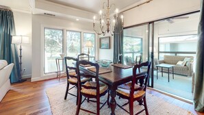 A dining area with a wooden table and chairs, a chandelier, and large windows. - Kiawah Exclusives | 4942 Turtle Point Villa (Kiawah Island)