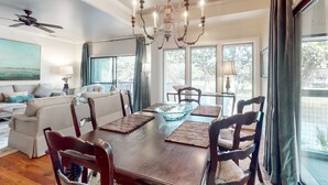A dining area with a wooden table, chairs, a chandelier, and large windows with curtains. - Kiawah Exclusives | 4942 Turtle Point Villa (Kiawah Island)