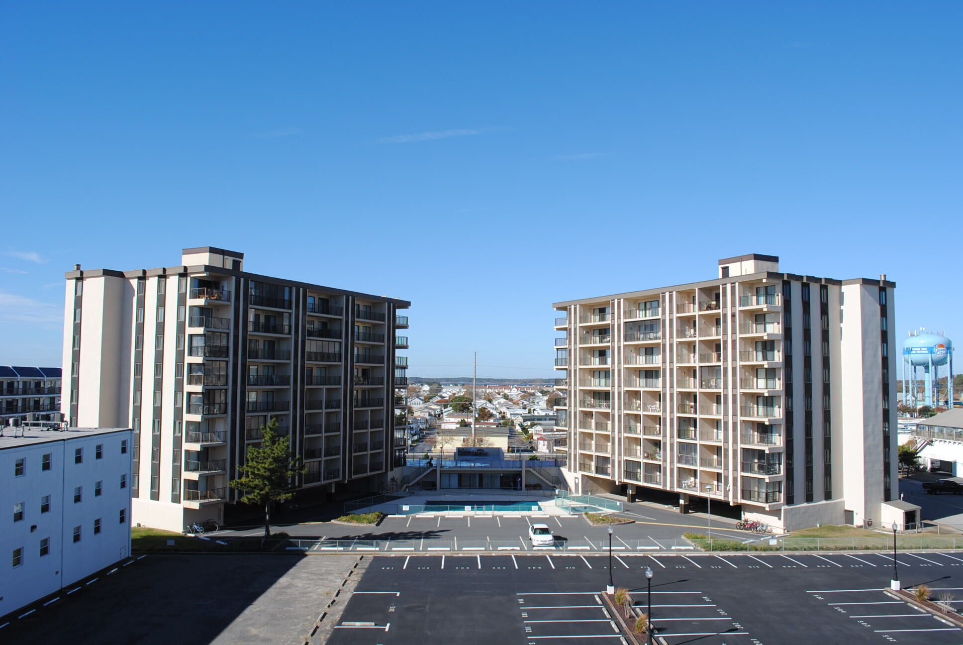 A modern apartment building with multiple floors and balconies, a parking lot in front, and a clear blue sky.