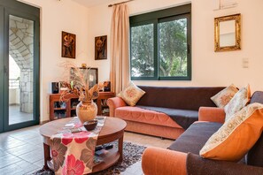 Ein Wohnzimmer mit einer Couch, einem Couchtisch und einem Fernseher. Ein Fenster bietet Blick auf Bäume.