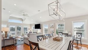 A spacious living room with a dining area, large windows, ceiling fan, and a flat-screen TV. - Heatable Pool, Short Walk to Beach, Luxury Living! Sweet Carolina (Folly Beach)