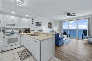 A modern kitchen with white cabinetry, a blue kettle, and a dining area with a blue sofa and a view of the ocean.