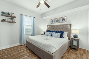 Ein Schlafzimmer mit einem großen Bett, einem Deckenventilator, einem Nachttisch mit Lampe und einem Fenster mit Vorhängen.