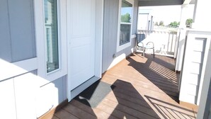 Een overdekt houten terras met een witte deur en ramen, dat uitkomt op een overdekte veranda.