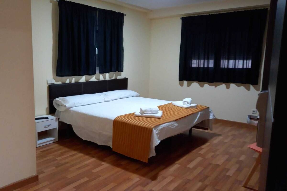 Ein ordentlich bezogenes Bett mit weißer Decke und brauner Wolldecke, ein Nachttisch mit Lampe und ein Fenster mit schwarzen Vorhängen.
