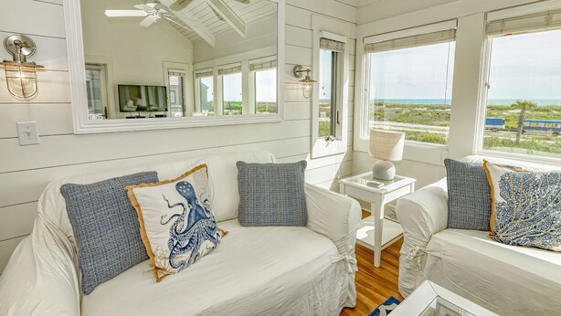 A white, coastal-style bedroom with a large mirror, a ceiling fan, and a view of the ocean.