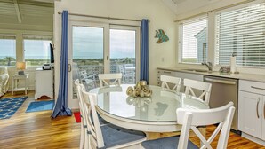 A spacious dining area with a glass table surrounded by white chairs, a ceiling fan, and a view of the ocean through large windows.