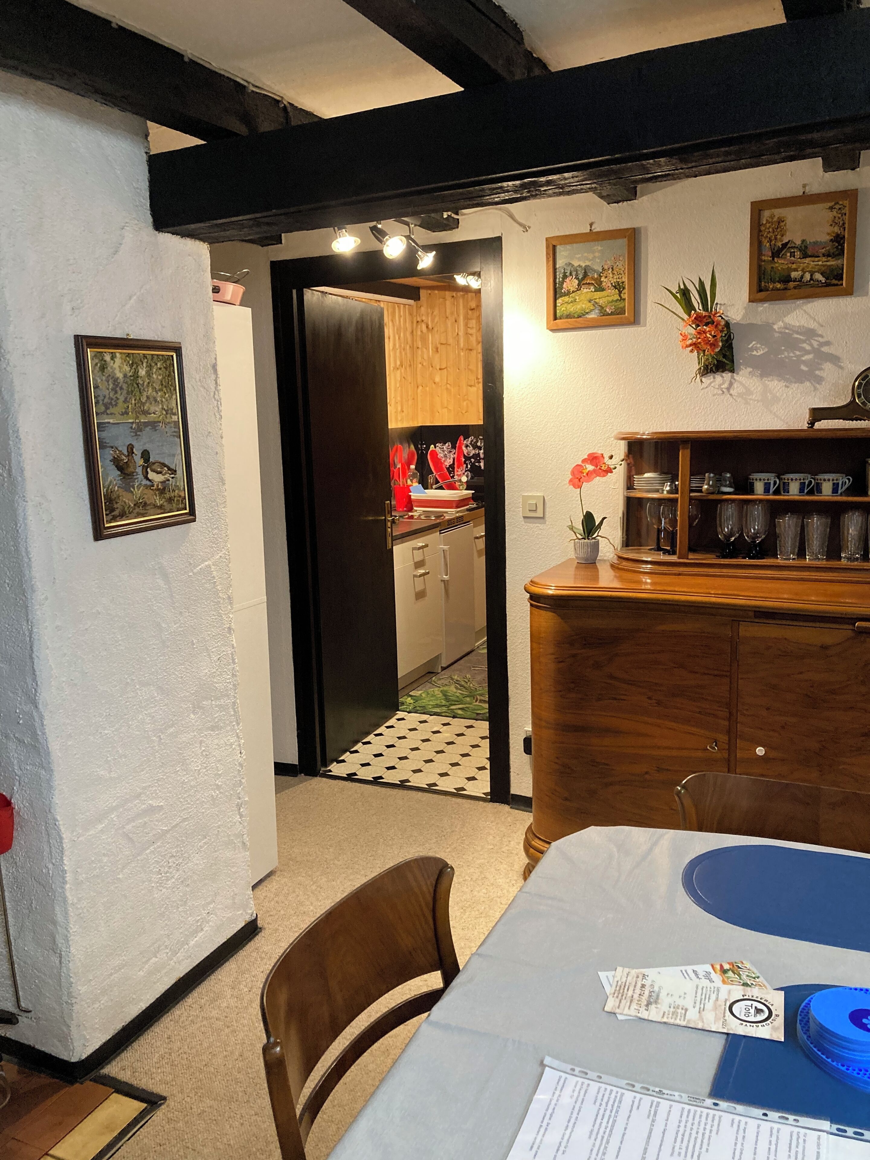A dining area with a table set for two, a wooden cabinet with glassware, and a door leading to a kitchen.