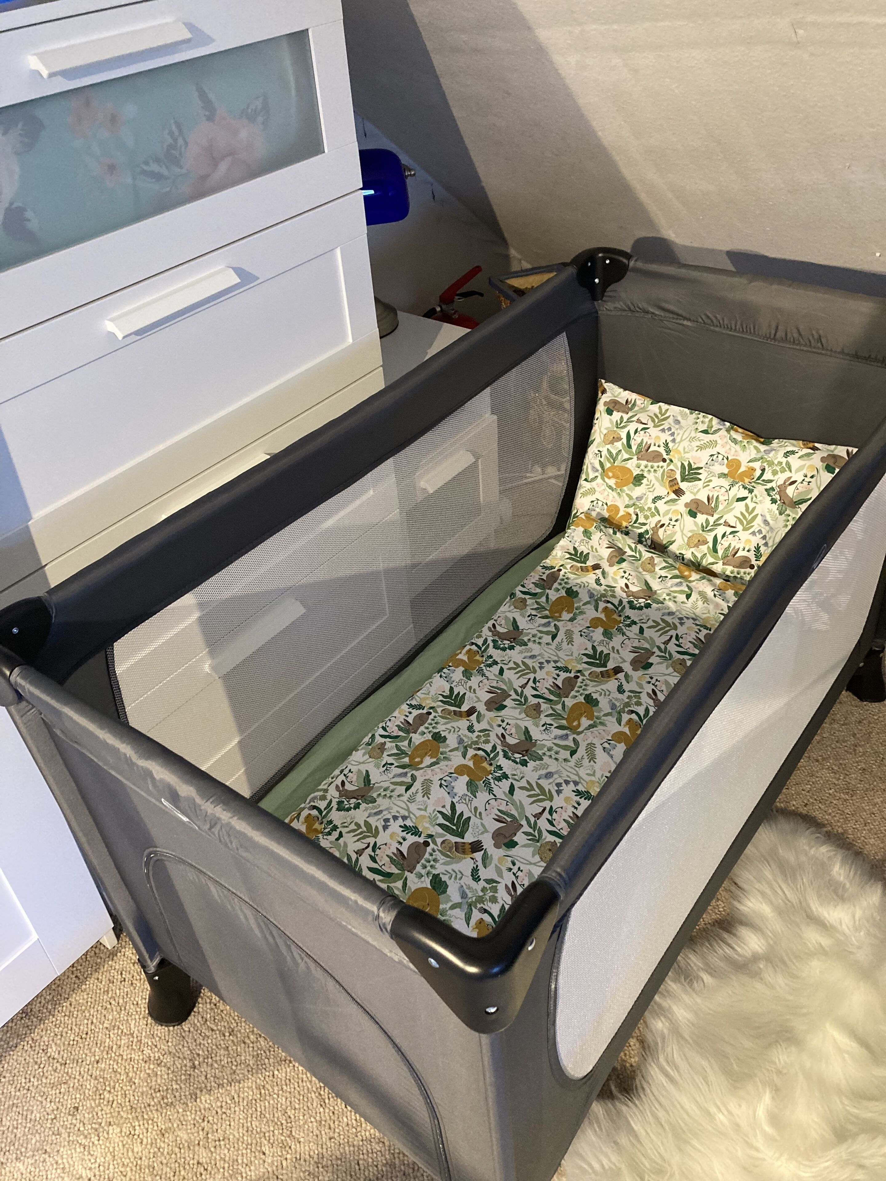 A portable crib with a mesh side and a floral-patterned mattress, placed in a room with a white cabinet and a blue container in the background.