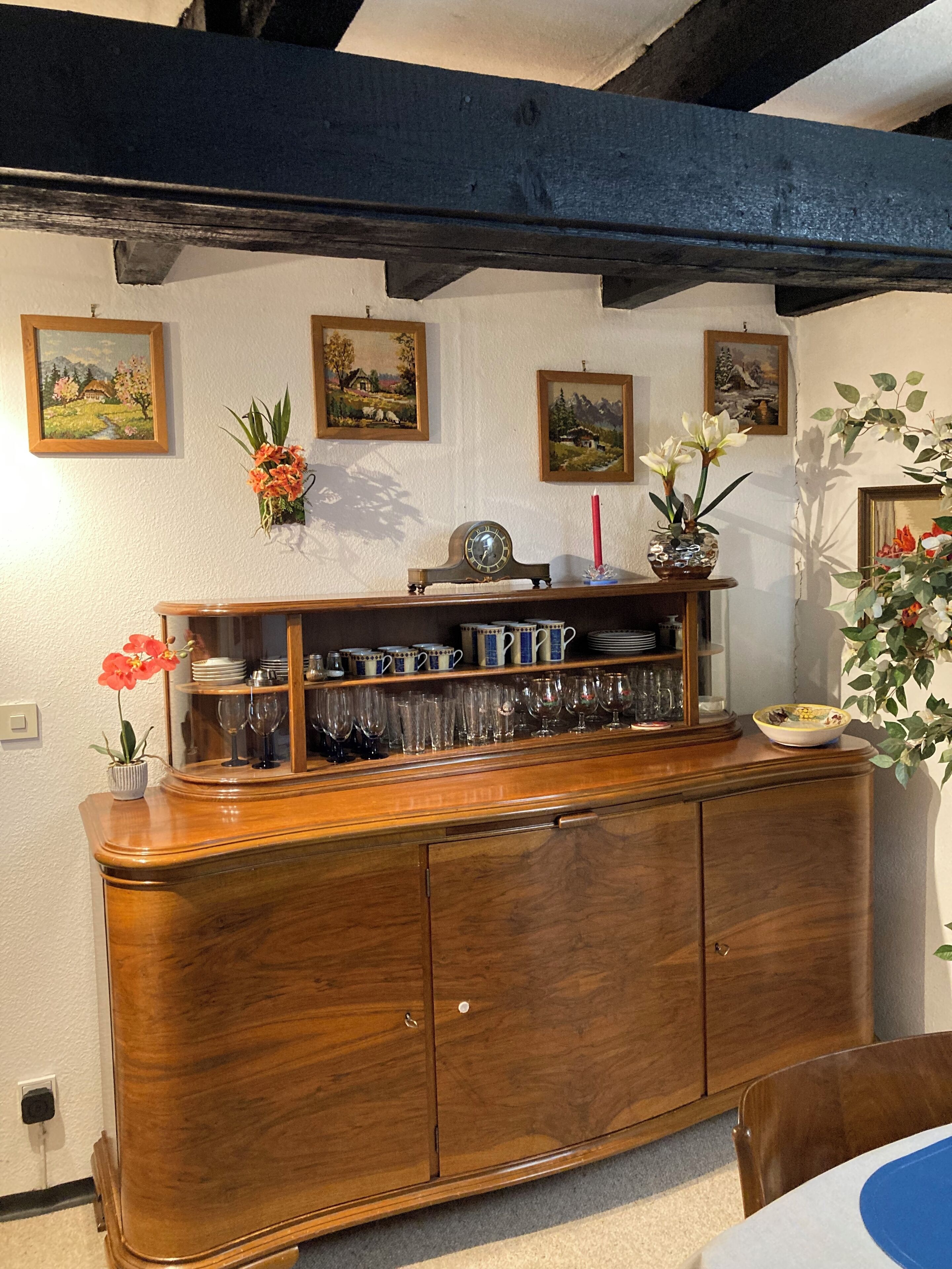 A wooden cabinet with glassware and dishes, paintings on the wall, and a vase with flowers.