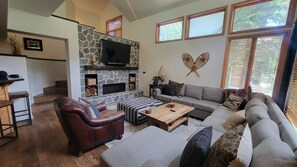 Un salon avec une cheminée en pierre, un canapé gris, un fauteuil en cuir marron et une table basse en bois.